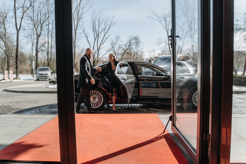 A couple stepping out of a luxury car onto a red carpet in an upscale outdoor setting.