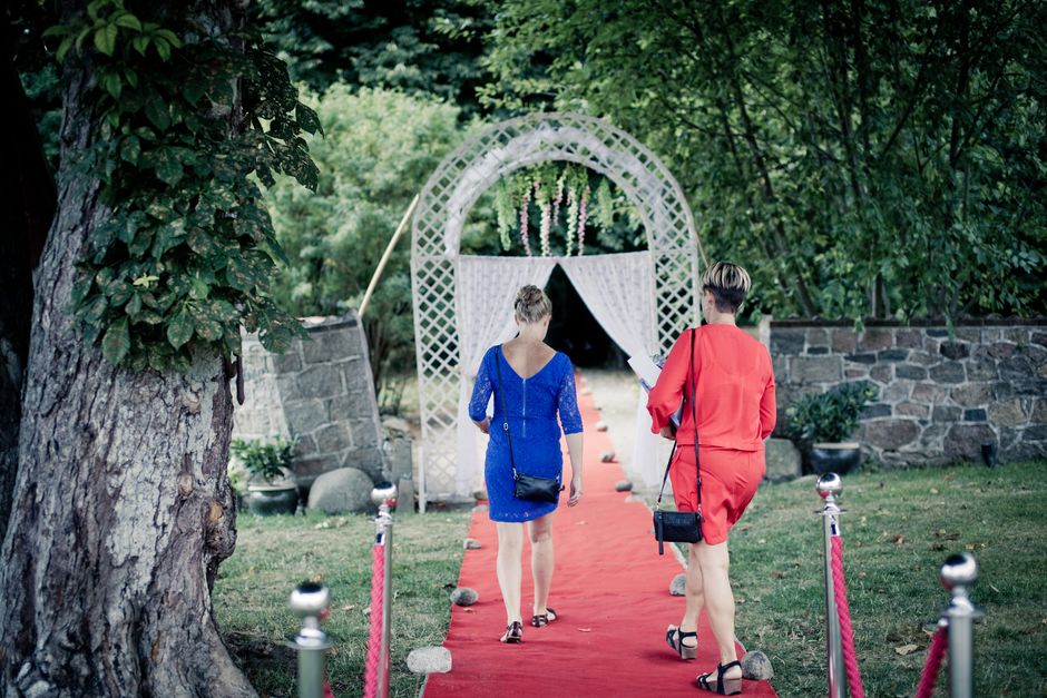 Two women walk on a red carpet through a garden towards a decorative archway.